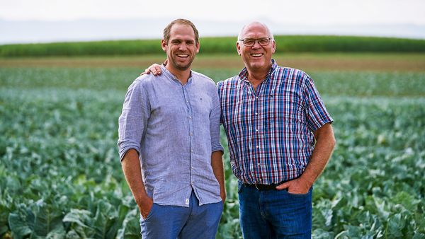 Zwei lächelnde Männer stehen auf einem Blumenkohlfeld, der Mann links trägt ein graues Hemd, der Mann rechts ein kariertes Hemd.