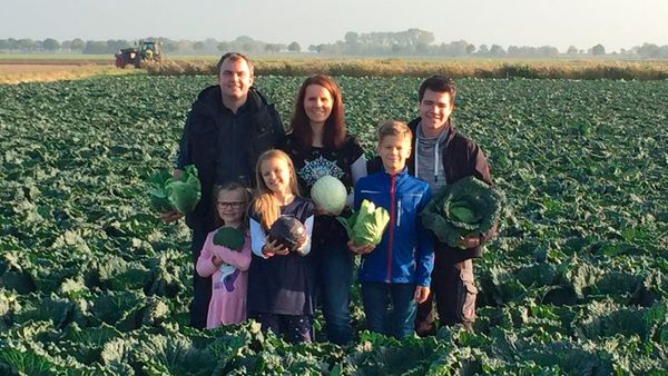 Familie mit drei Kindern steht in einem großen Kohlfeld und hält verschiedene Kohlarten aus Eigenproduktion. Im Hintergrund sind weitere Felder und ein Traktor zu sehen.