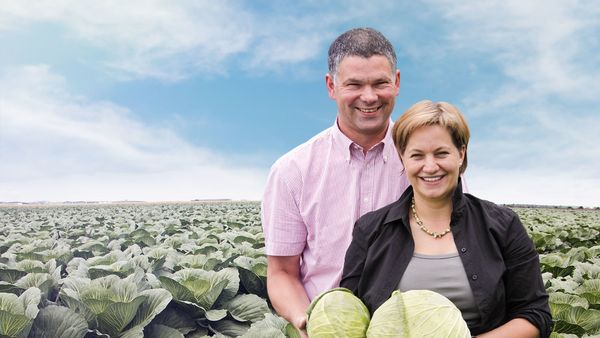 Lächelnder Mann und Frau stehen in einem weiten Kohlfeld unter blauem Himmel, halten zwei frisch geerntete Kohlköpfe aus Eigenproduktion.