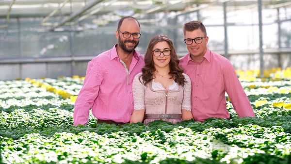 Ein Mann, eine Frau und ein junger Mann in traditionellen bayerischen Kleidern stehen in einem Gewächshaus voller grüner Pflanzen.