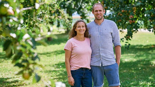 Lächelnder Mann in grauem Hemd und Frau in rosa Shirt stehen im grünen Obstgarten mit Obstbäumen im Vorder- und Hintergrund.