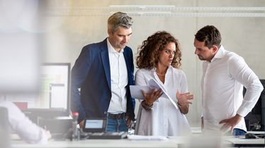 Drei Personen, zwei Männer und eine Frau, diskutieren in einem Büro und halten Dokumente, sprechen über die Vermarktung im Obst und Gemüsehandel.