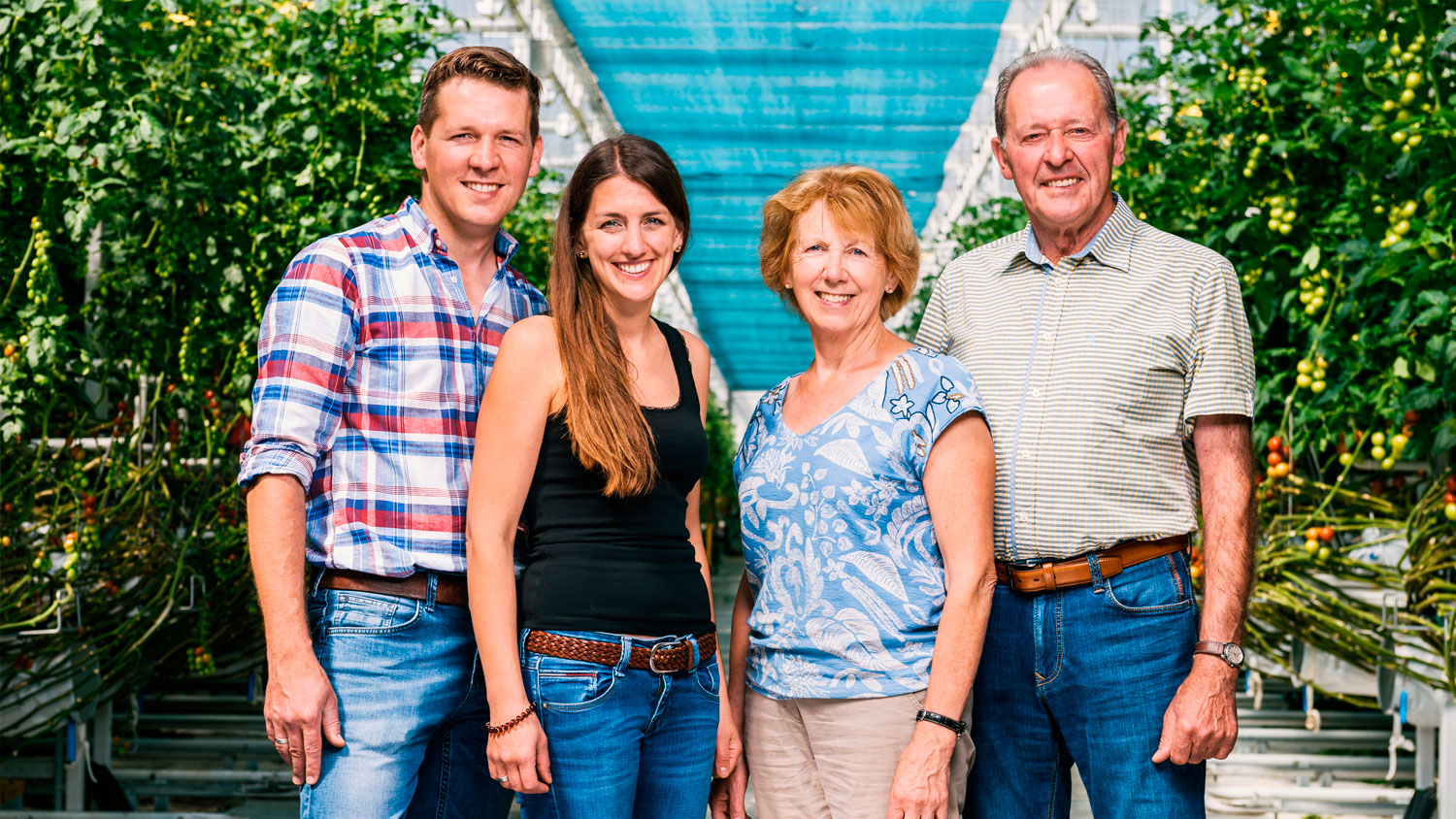 Vierköpfige Familie, zwei Männer und zwei Frauen, stehen in einem Gewächshaus mit grünen Tomatenpflanzen aus Eigenproduktion im Hintergrund.