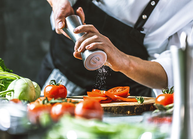 Ein Koch in Schürze würzt geschnittene Tomaten auf einem Holzbrett, frische Tomaten und Gemüse vom Foodservice im Vordergrund.