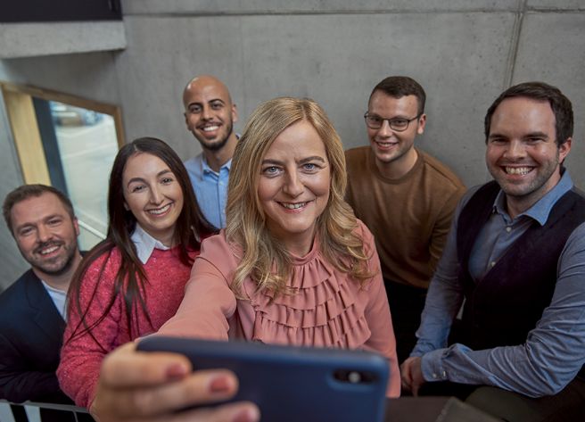 Sechs lächelnde Personen in Bürokleidung posieren für ein Selfie. Die Frau in der Mitte hält das Smartphone.